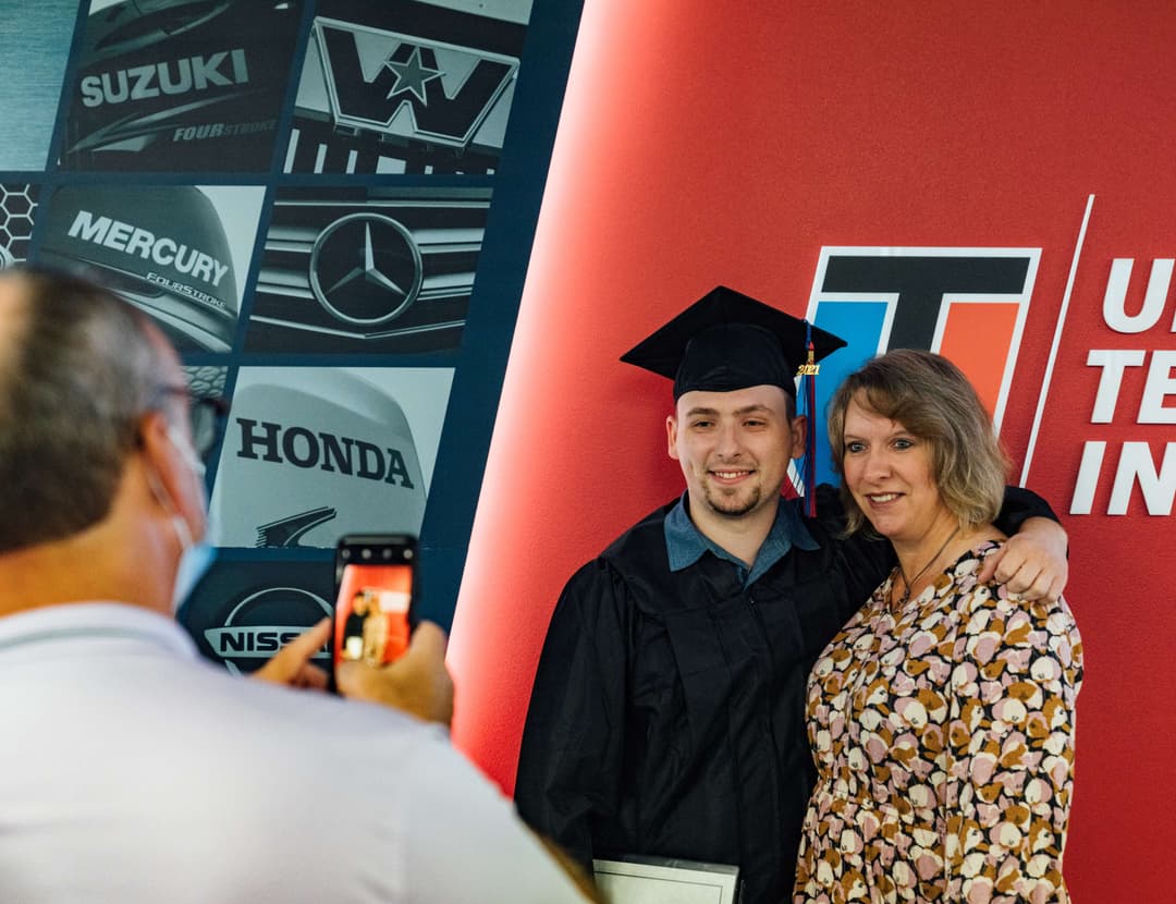 UTI trade school graduate wearing a black cap and gown with honor cords, smiling at commencement ceremony.